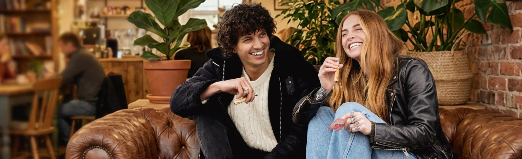 Two people sitting on a couch in a cozy cafe setting with plants and wooden furniture.